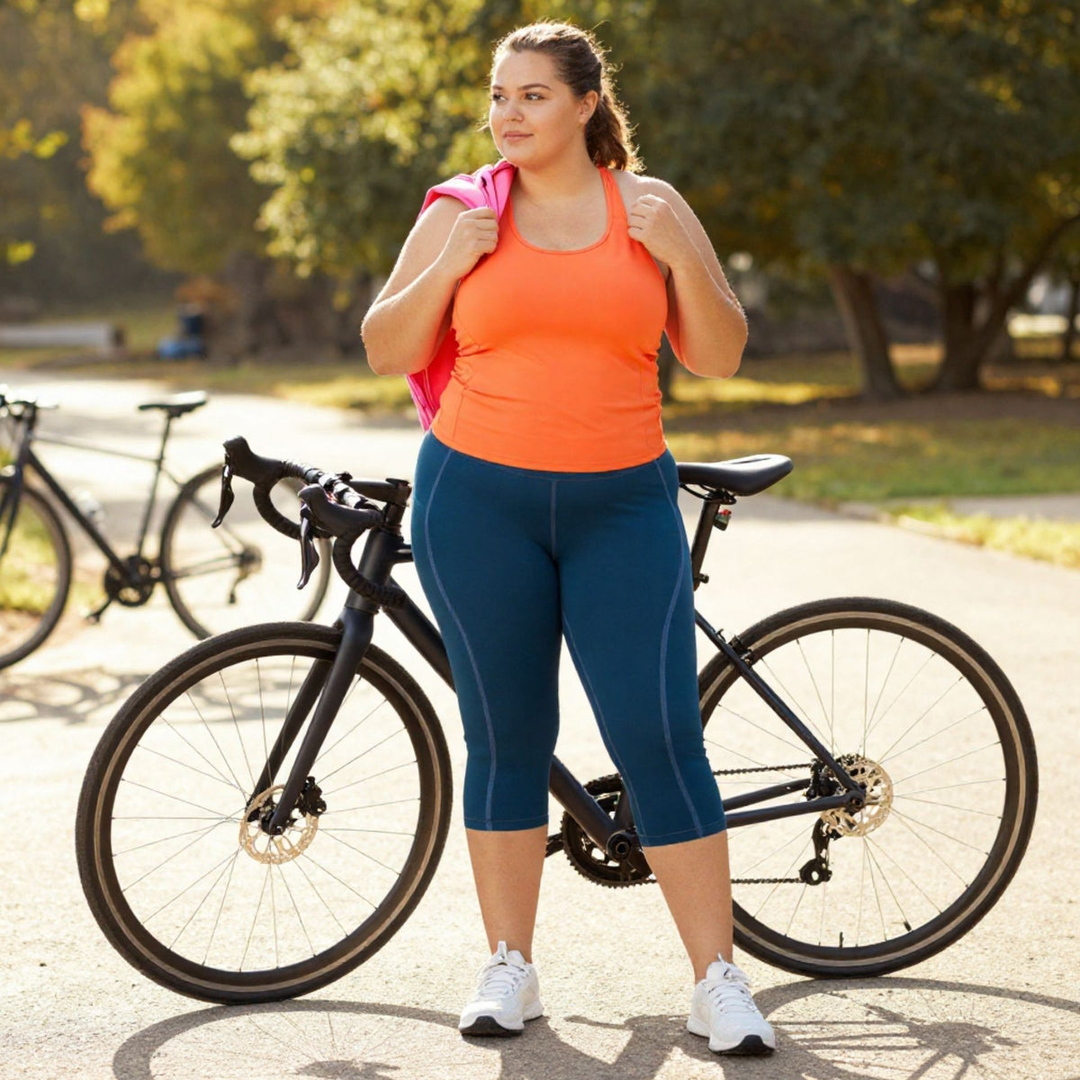 A woman in an orange tank top and teal capri leggings stands next to a black road bike on a paved path in a sunny park, holding a pink bag over her shoulder.