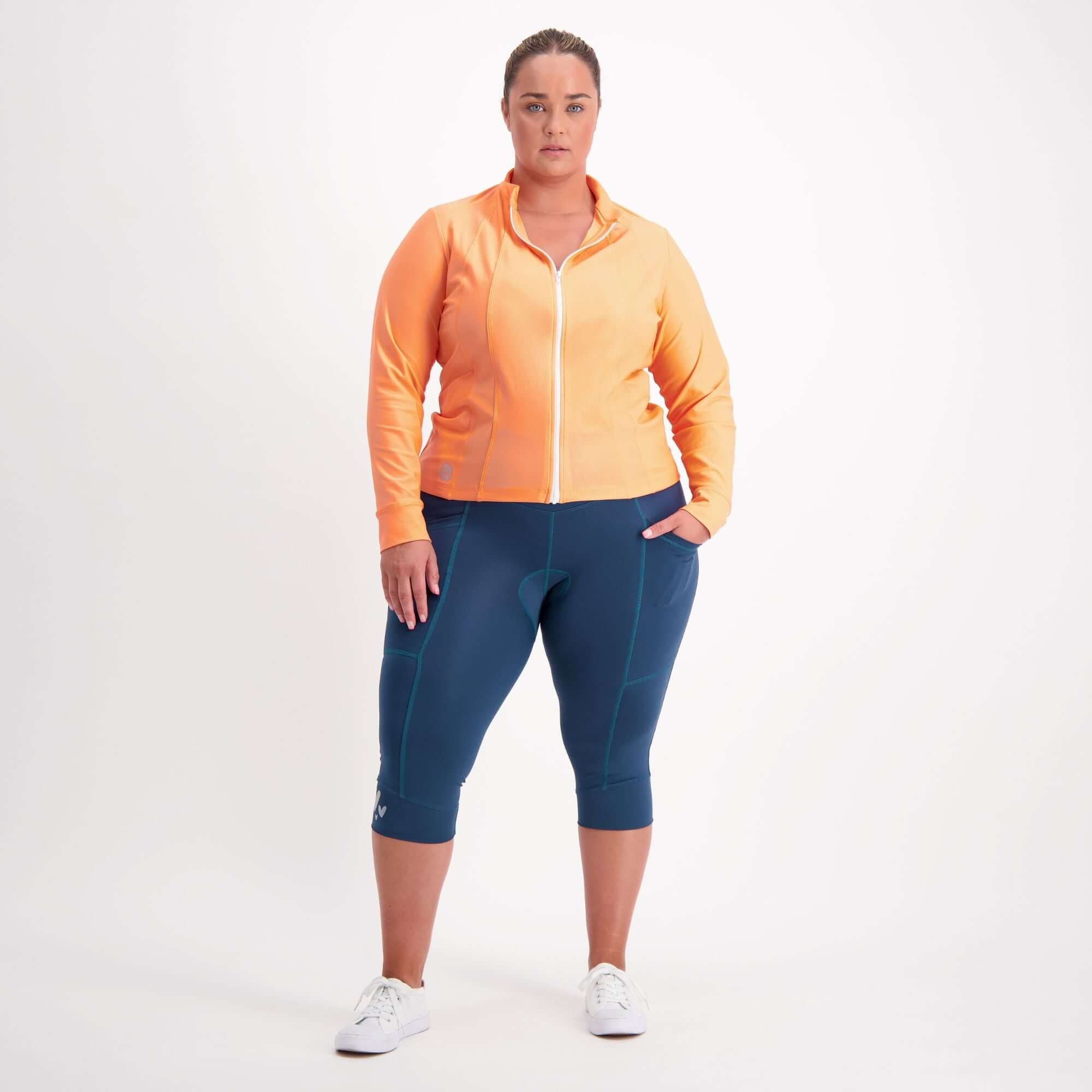 Woman wearing an orange jacket and forest green padded three quarter bike pants on a white background