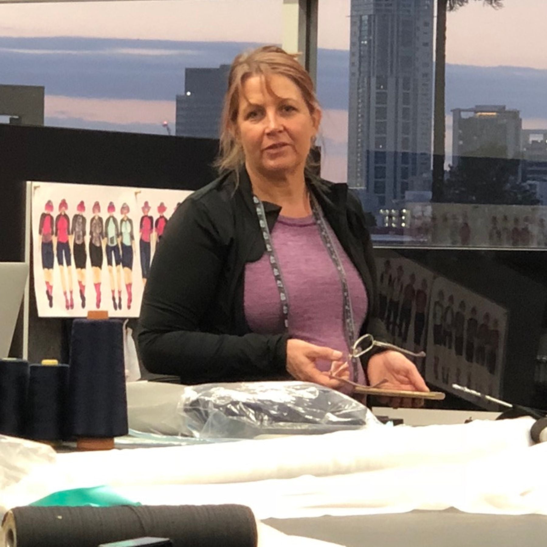 A woman in a purple shirt and black cardigan stands behind a sewing table with fabric spools and scissors, with a fashion design poster and a city skyline visible in the background.