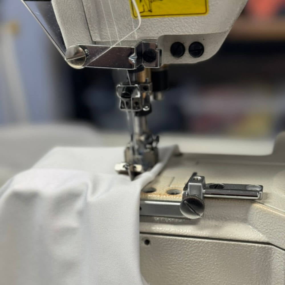 Close-up of a sewing machine in use with white fabric under the press foot, showing the needle area, presser foot, and part of the machine body.