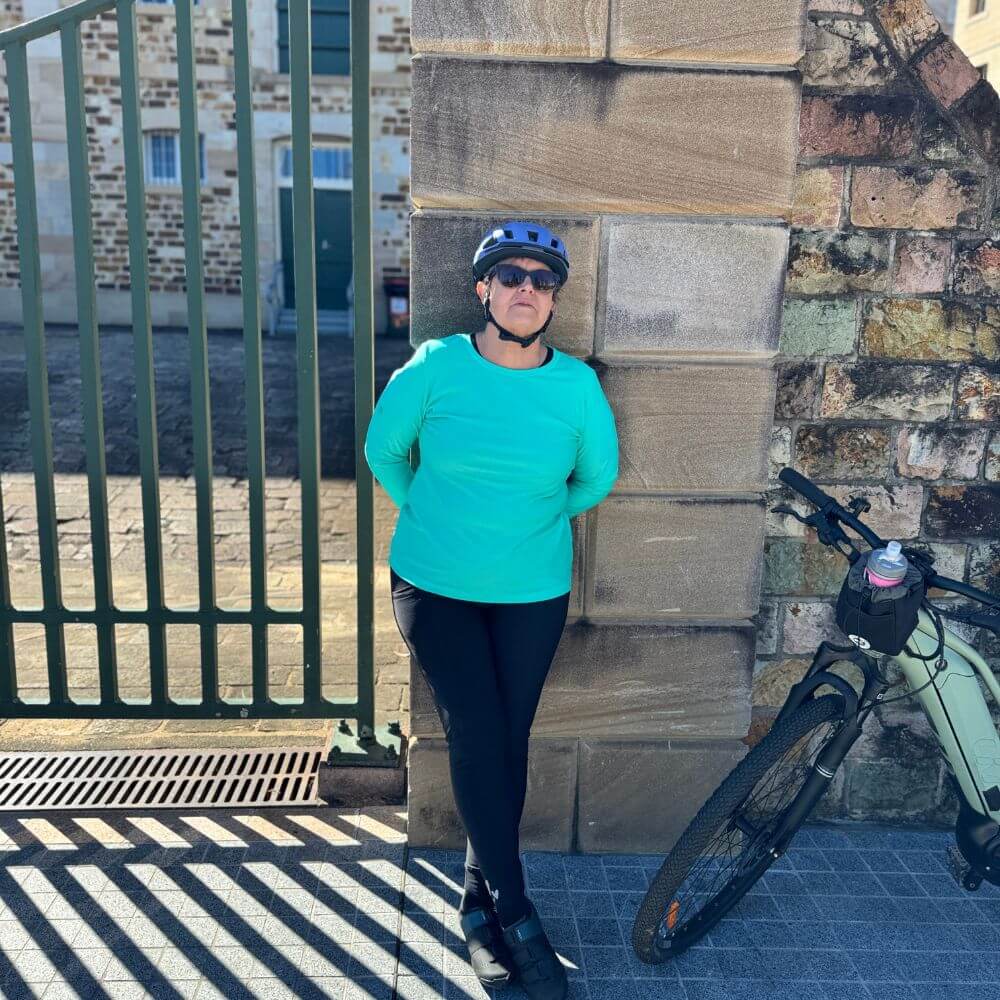 Person in teal shirt and black helmet standing next to a bicycle against a stone wall.