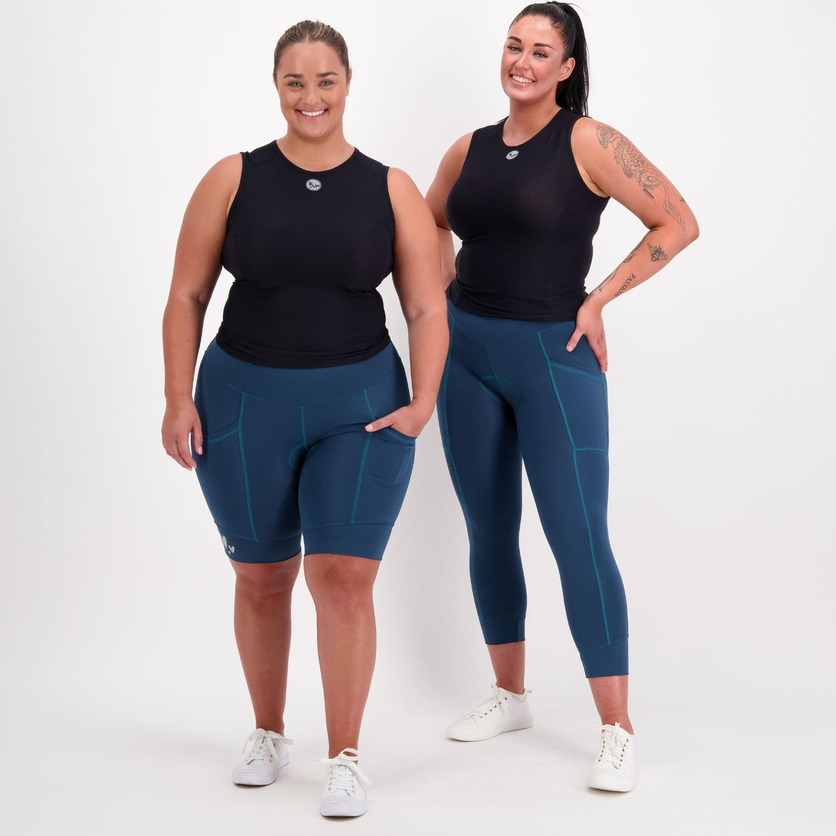 Two women modeling matching athletic wear against a plain light gray studio background. The set includes a sleeveless black top and fitted leggings or biker shorts in a dark teal-blue color with subtle lighter blue seam lines.