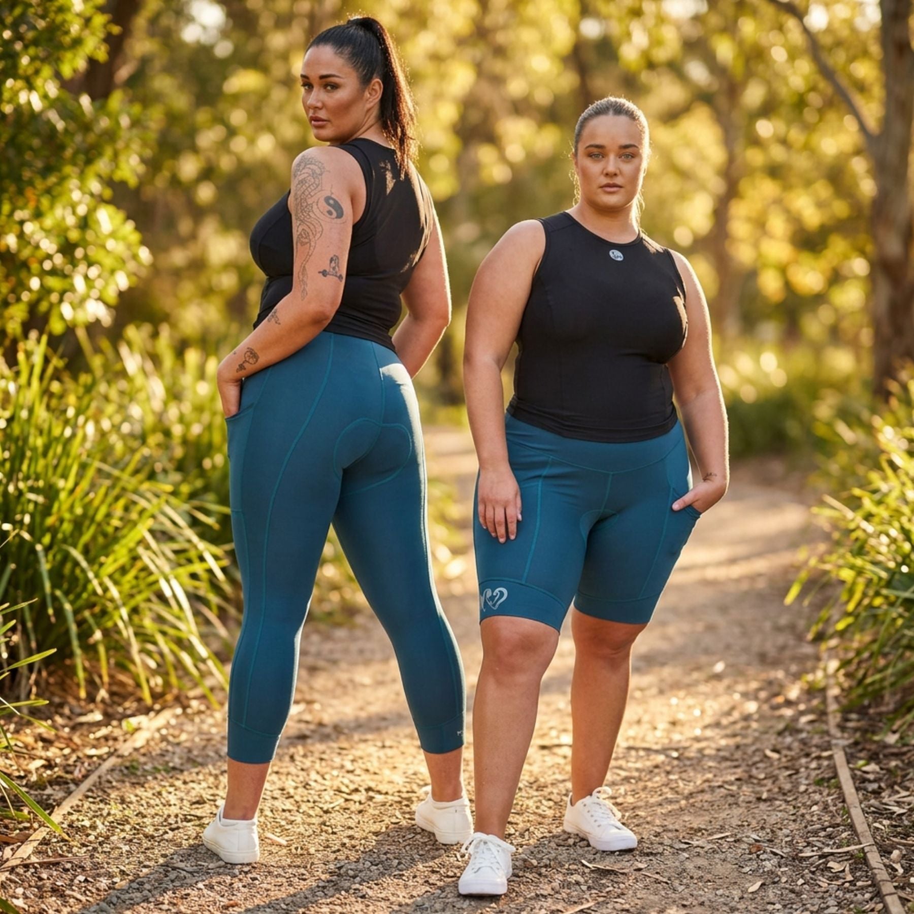 Two women modeling athletic wear on a sunlit forest path. The woman on the left wears a black sleeveless top and teal full-length leggings, while the woman on the right wears a black sleeveless top and matching teal fitted shorts.