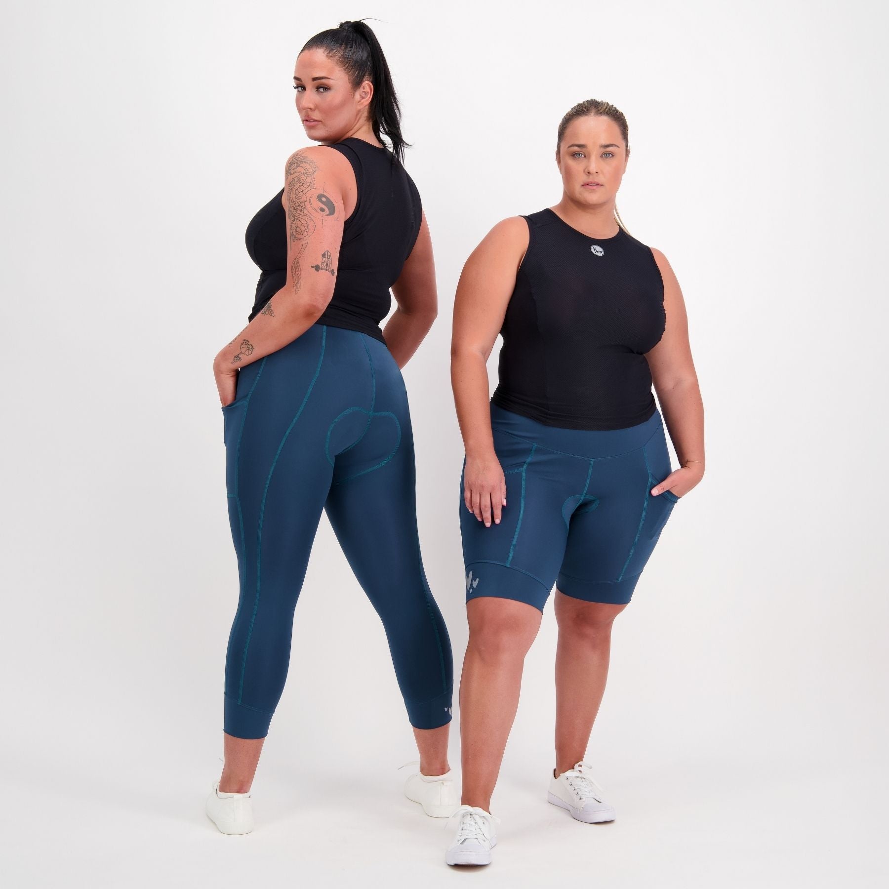 Two women modeling dark teal athletic leggings and matching fitted shorts with a black sleeveless top, standing against a plain light gray studio background.