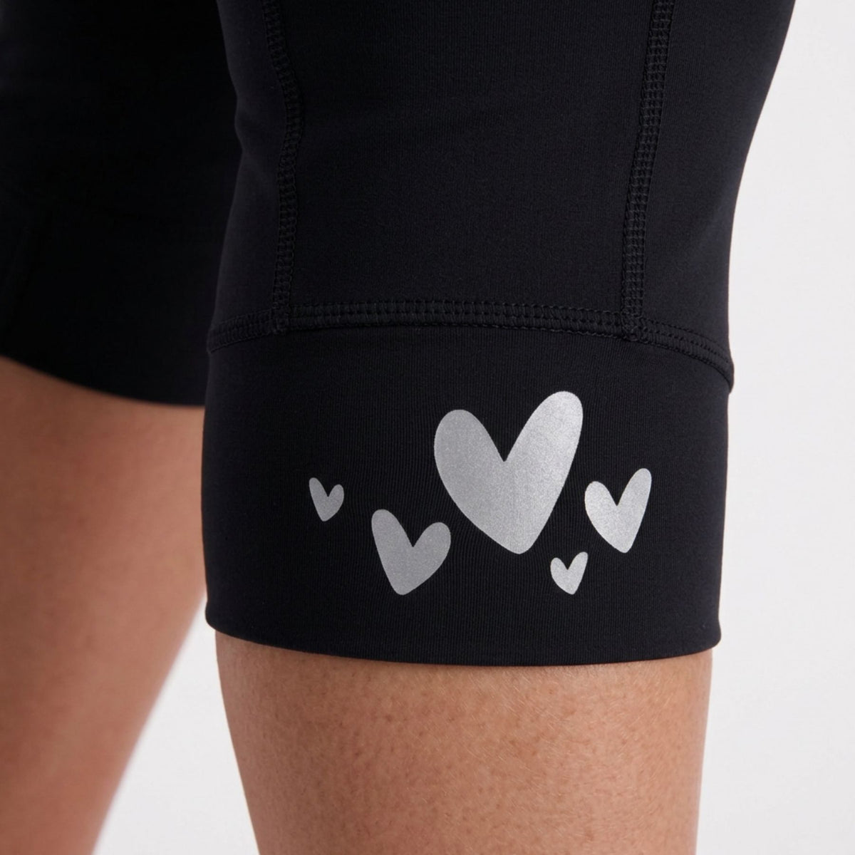 Close-up of black padded bike shorts worn on a person's leg, with a cuff featuring a silver heart graphic and small matching heart shapes on a light gray background.