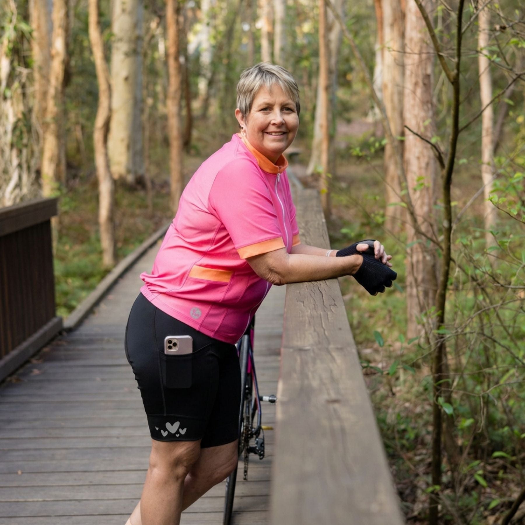 A woman wearing a pink cycling jersey and black shorts leans on a wooden railing on a boardwalk surrounded by trees. A bicycle is partially visible beside her, and a phone is tucked into her shorts.