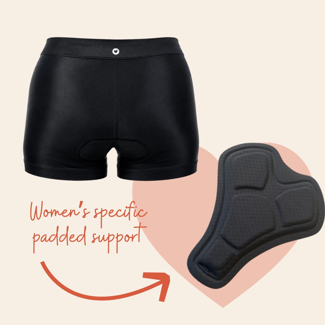 Black women's cycling shorts with a padded support insert, shown on a light beige background with a pink heart shape and orange text reading 'Women's specific padded support.'.