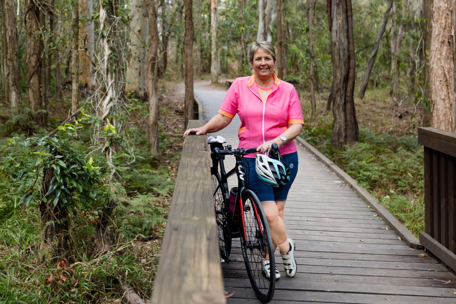 female cyclist in pink jersey walking a bicycle along a wooden bridge