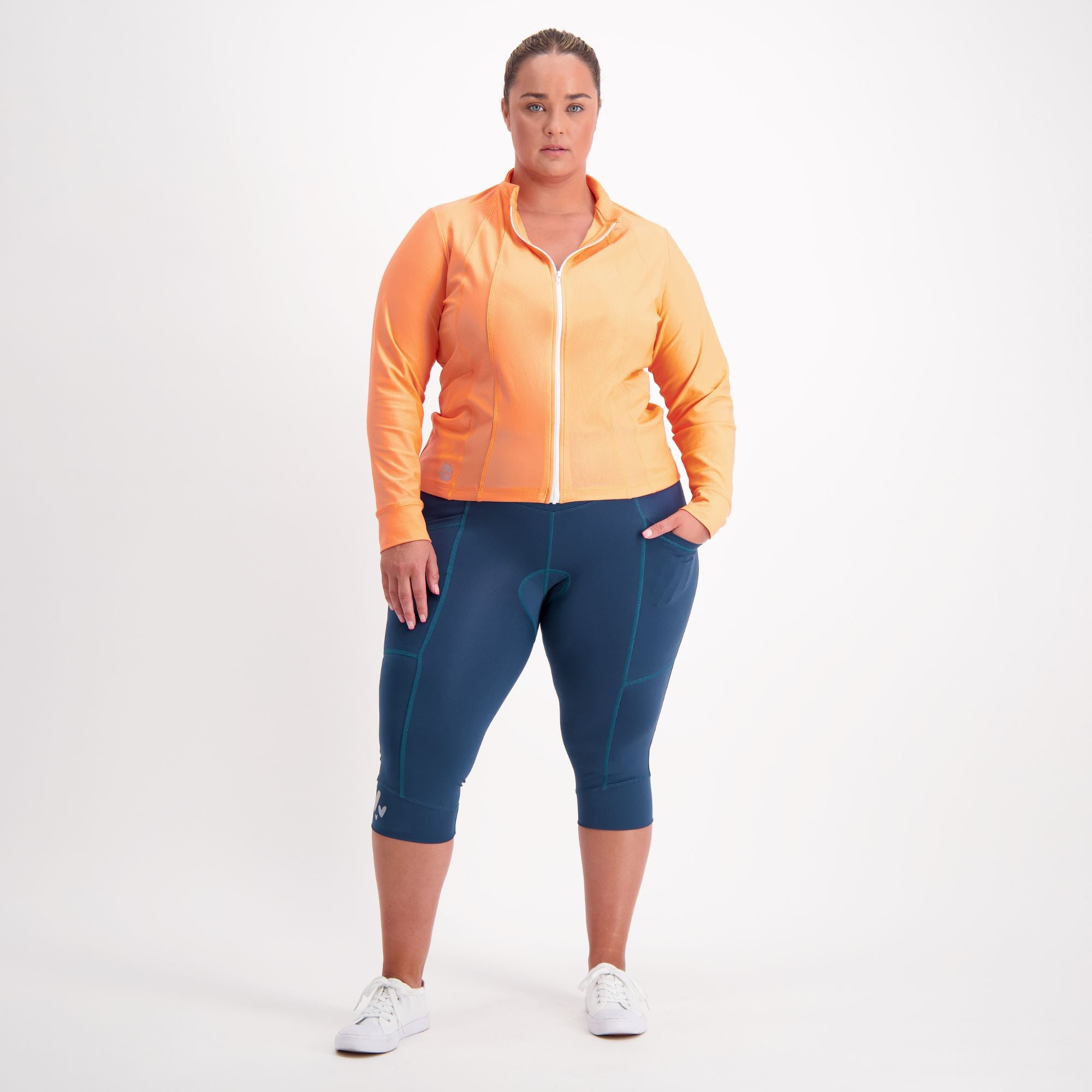 Woman wearing an orange jacket and forest green padded three quarter bike pants  on a white background