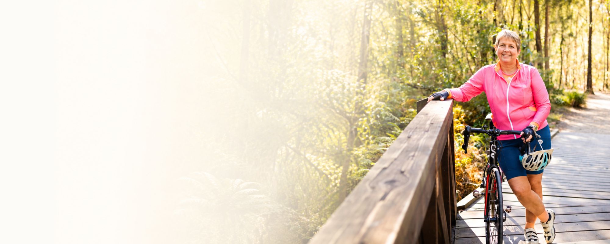 A woman in a pink zip-up jacket and dark shorts stands on a wooden boardwalk beside a bicycle, holding a helmet and smiling in a sunlit forest setting.