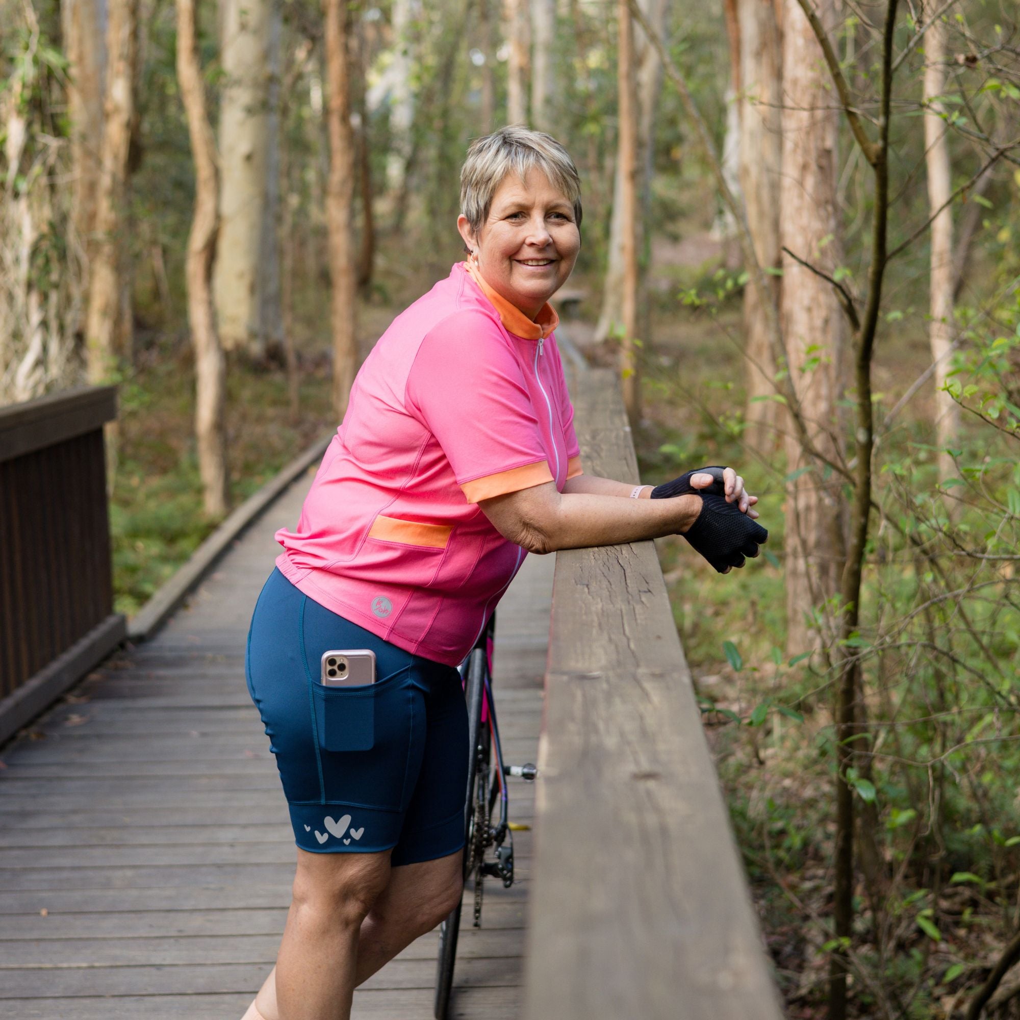 Woman in pink ccyling jersey and forest green padded bike shorts standing on a wooden path in a forest
