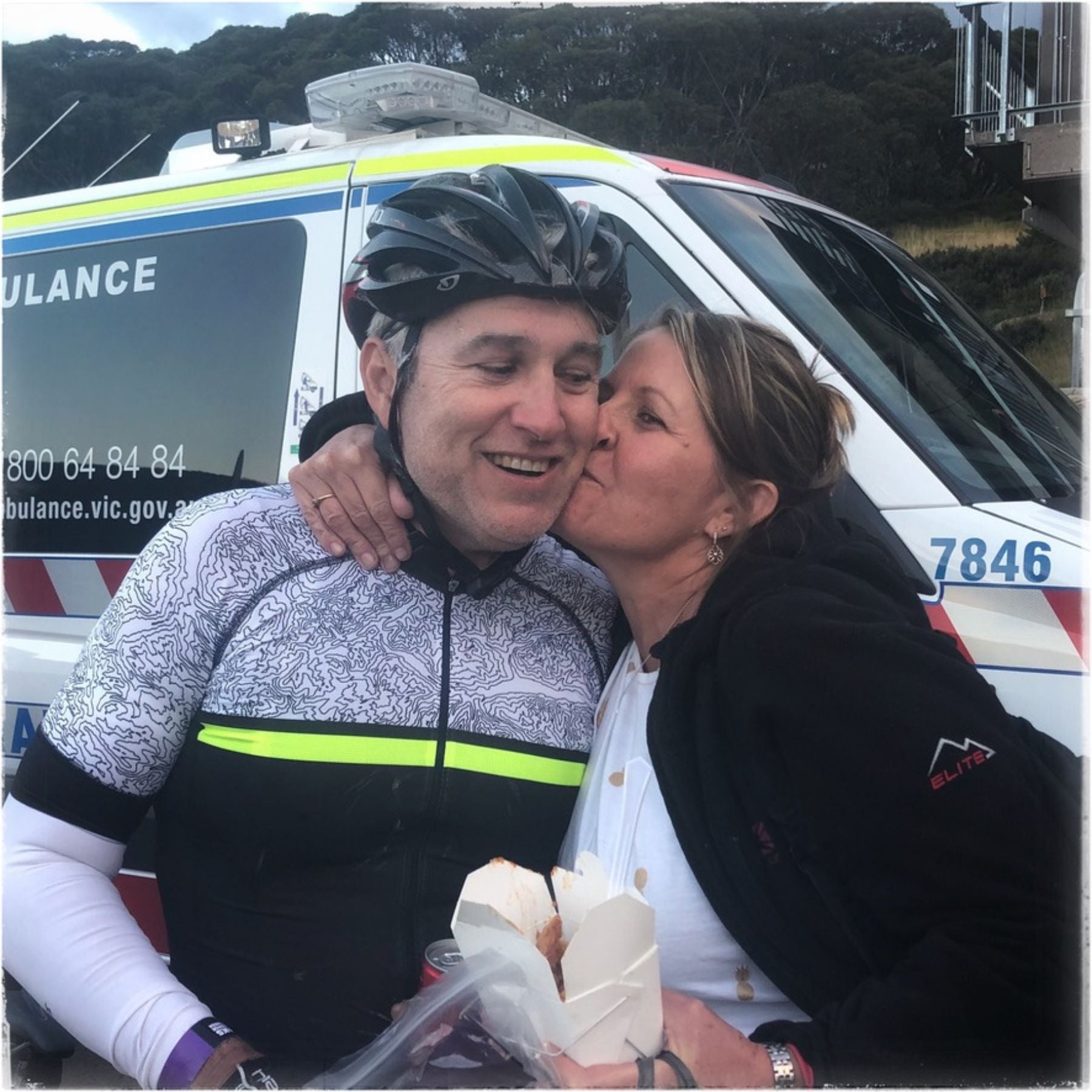 A smiling cyclist wearing a black helmet and a black-and-white patterned cycling jersey kisses a woman on the cheek in front of an ambulance. The cyclist is holding a food box in one hand.