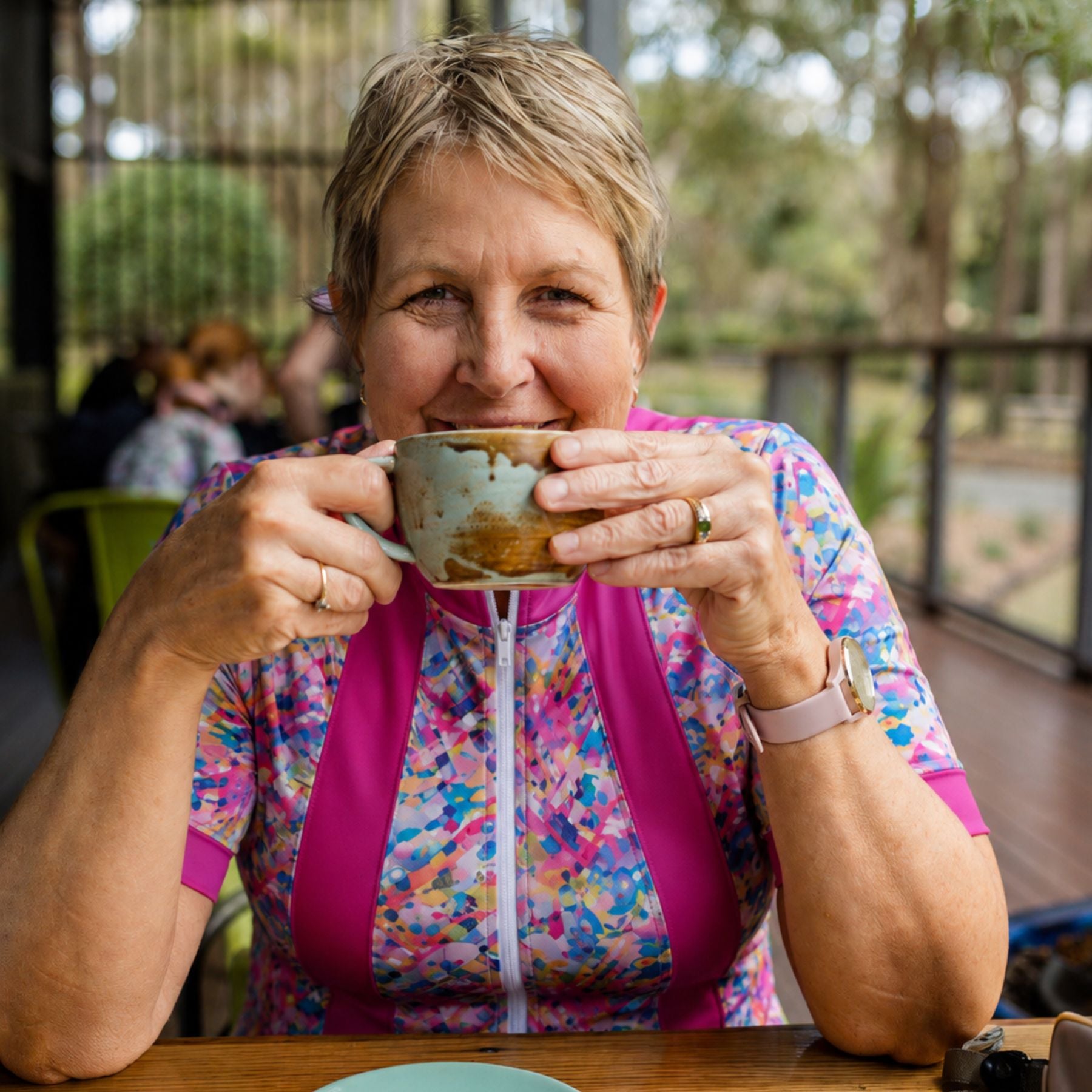 A smiling woman with short blonde hair holds a ceramic mug to her lips while wearing a colorful floral zip-up top with bright pink side panels, seated at a wooden table in an outdoor patio setting.