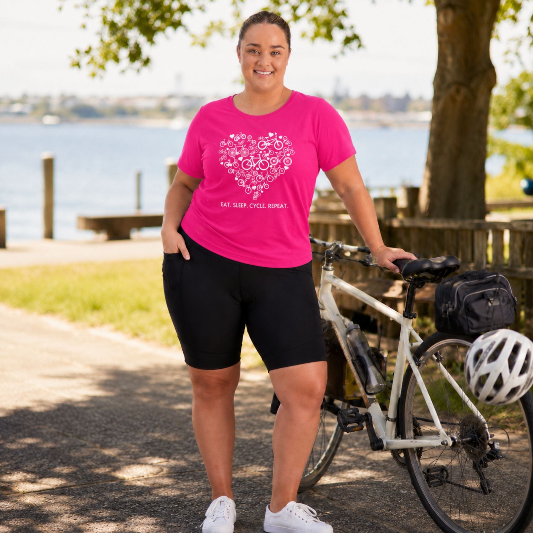 A woman stands outdoors beside a bicycle on a park path, wearing a bright pink T-shirt with a white floral heart graphic and the text 'I'M NOT AN ANIMAL SHIT.' She is also wearing black fitted shorts and white sneakers.