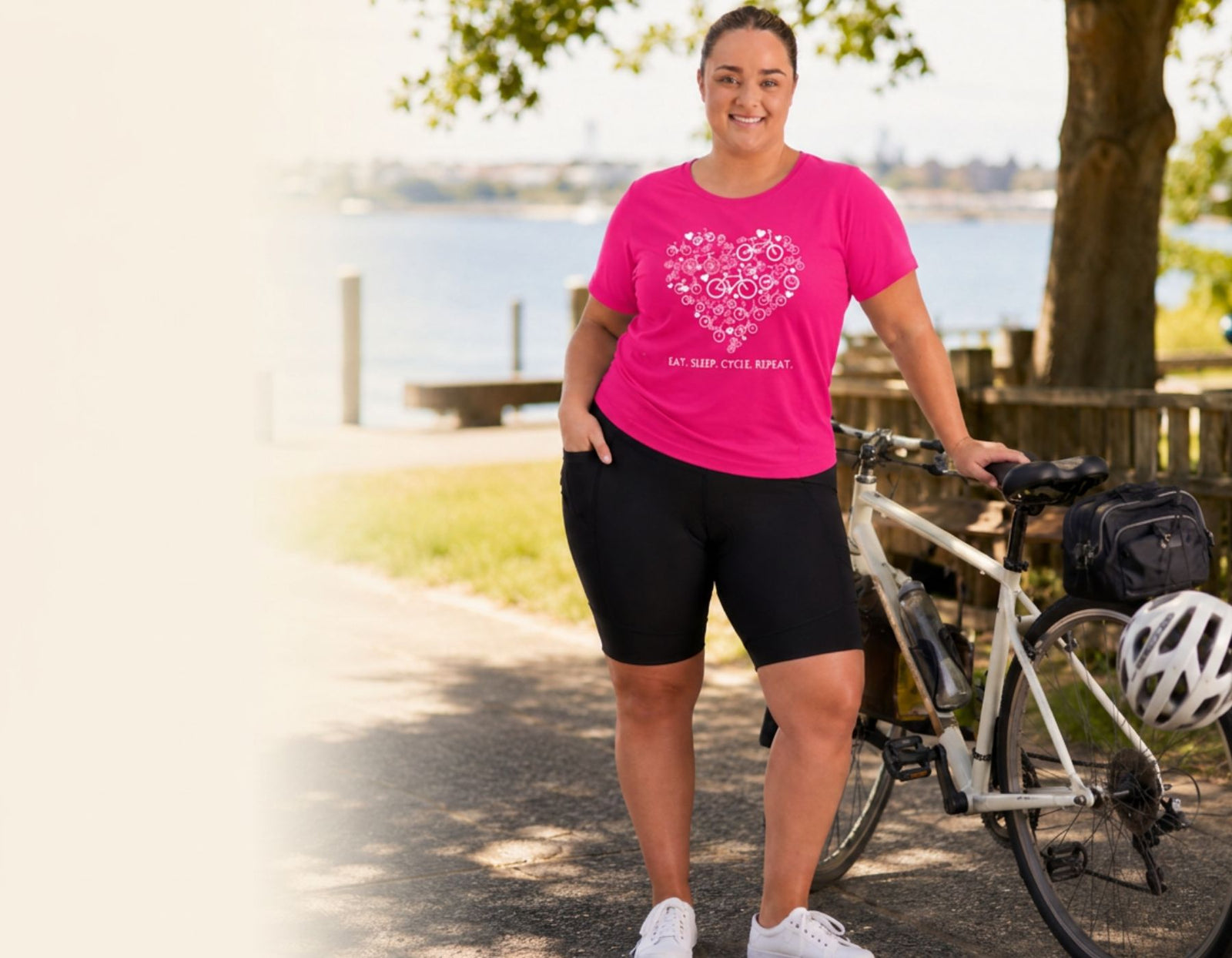 A woman stands outdoors next to a bicycle on a path near water, wearing a bright pink graphic T-shirt and black cycling shorts. The shirt has a white floral heart design with the text 'EAT SPORTY STYLES' underneath.