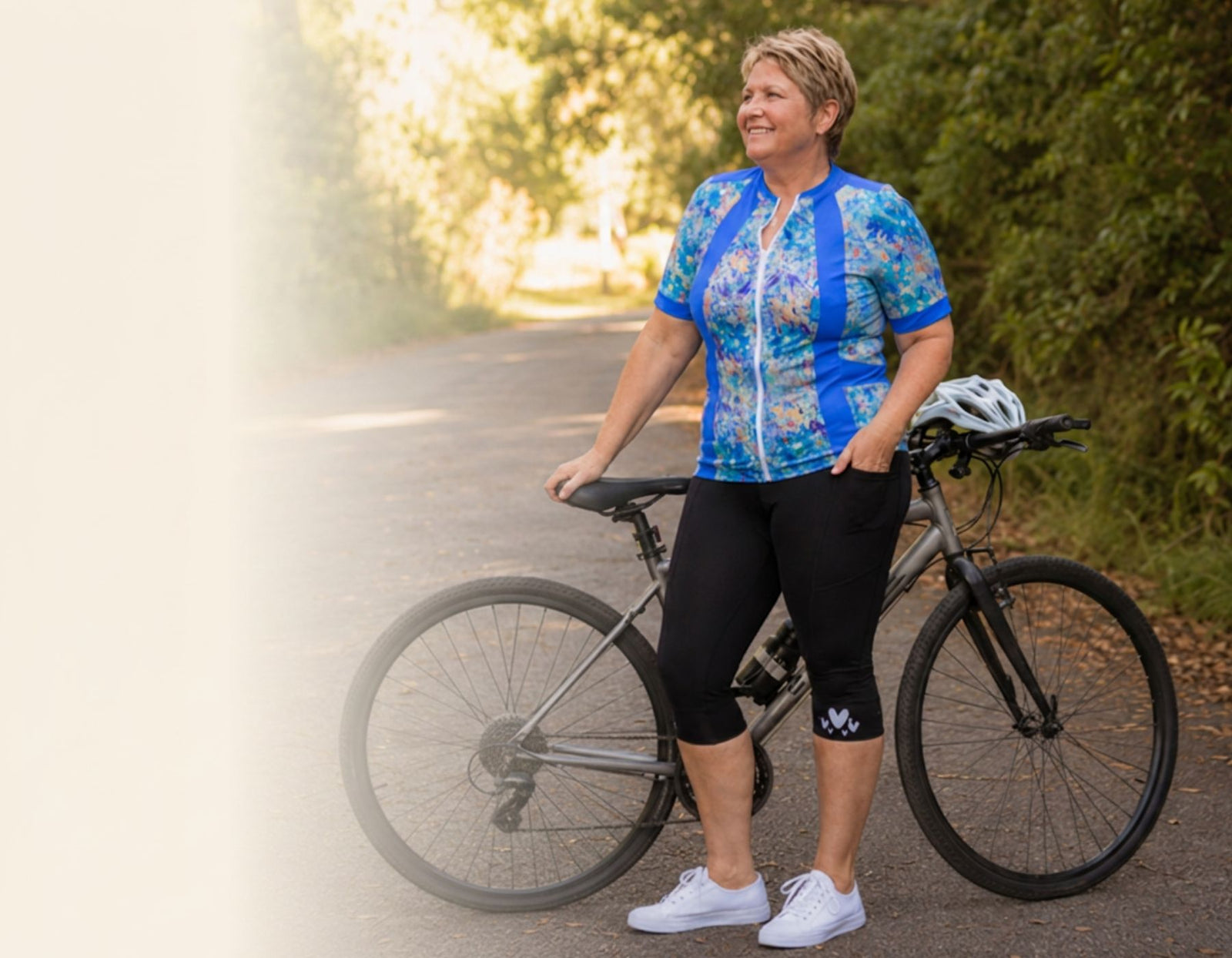A woman stands on a paved path beside a bicycle, wearing a blue and white patterned cycling jersey, black capri leggings, and white sneakers. A white helmet rests on the bicycle frame.
