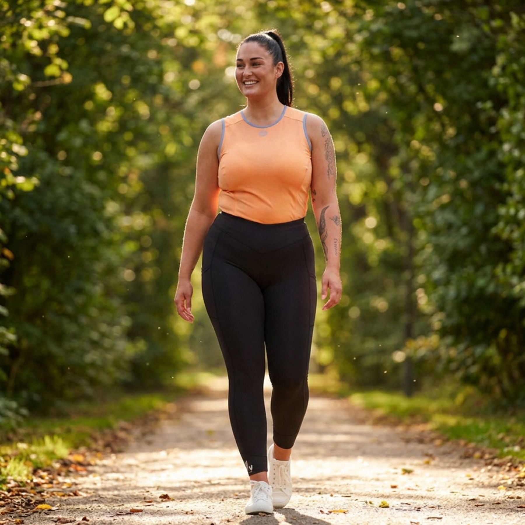 A woman walking on a sunlit dirt path surrounded by green trees, wearing an orange sleeveless athletic top and black leggings with white sneakers.