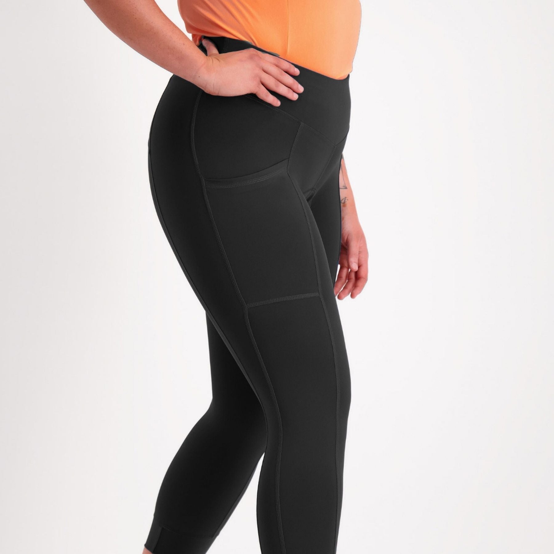 A side-profile view of a person wearing black high-waisted leggings with a peach-colored top, cropped from the waist down against a light gray studio background.