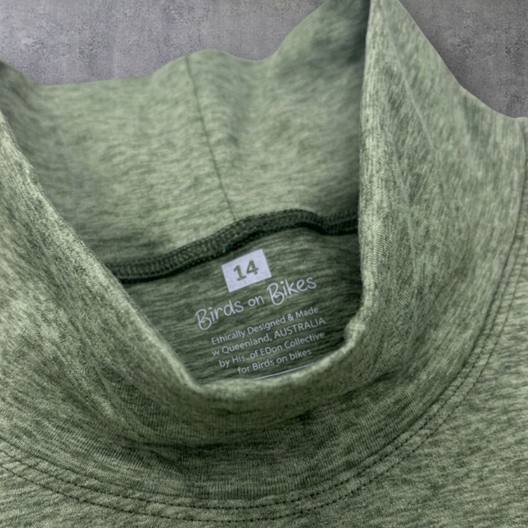 Close-up of a heathered sage green athletic turtleneck collar with a visible size tag reading 14 and the brand name Birds on Bikes printed inside the neck.