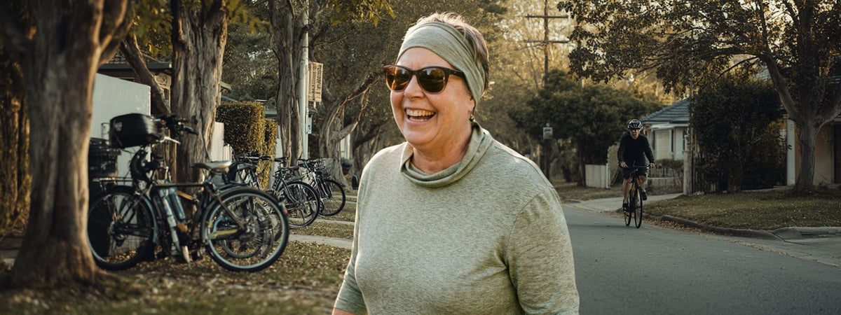 A smiling woman wearing a light green knit turtleneck, a matching green headband, and sunglasses stands on a tree-lined street with bicycles parked on the left. Another cyclist is visible in the background.